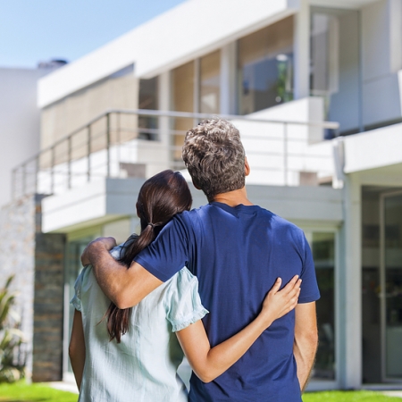 couple embracing in front of new big modern house, outdoor rear view back looking at their dream home